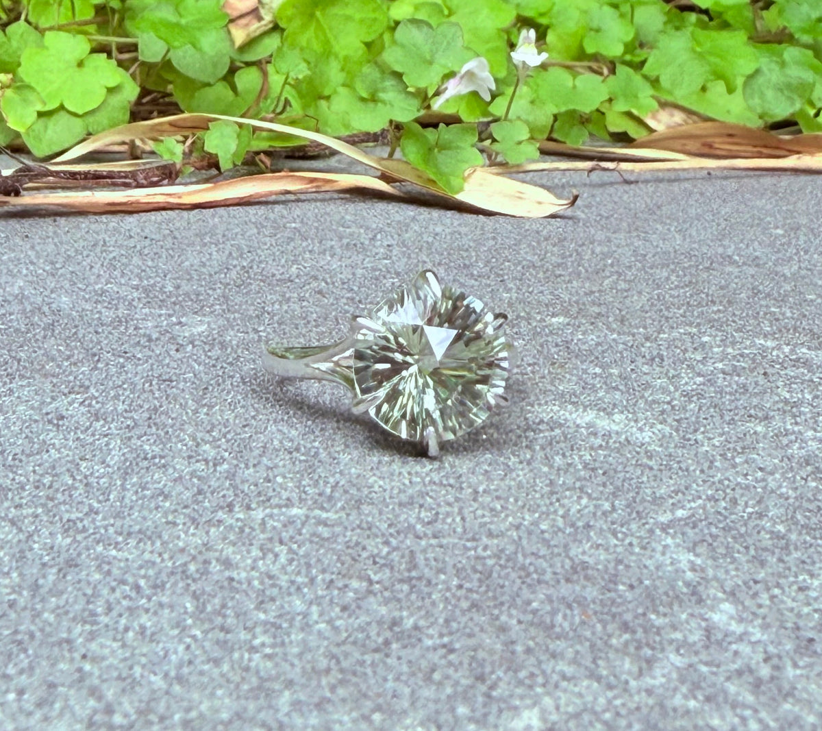 A single round brilliant-cut green amethyst ring set in silver-toned metal, resting on a gray concrete surface with green leafy plants in the background.
