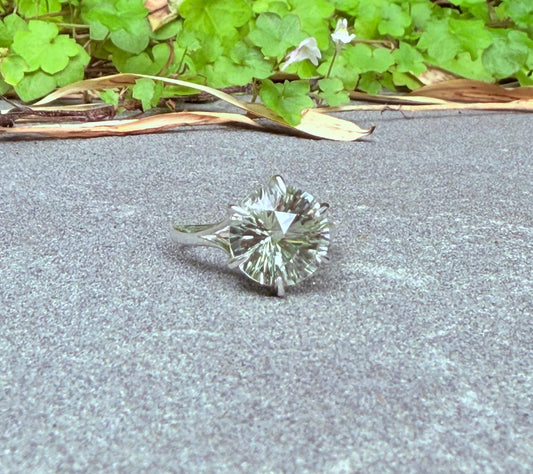 A single round brilliant-cut green amethyst ring set in silver-toned metal, resting on a gray concrete surface with green leafy plants in the background.