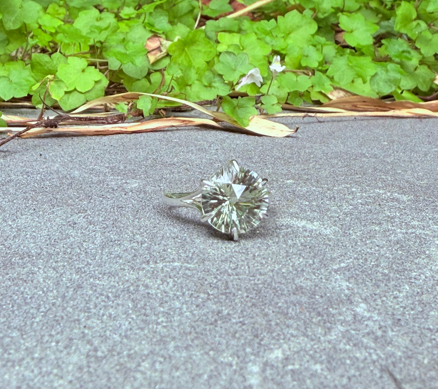 A single round brilliant-cut green amethyst ring set in silver-toned metal, resting on a gray concrete surface with green leafy plants in the background.