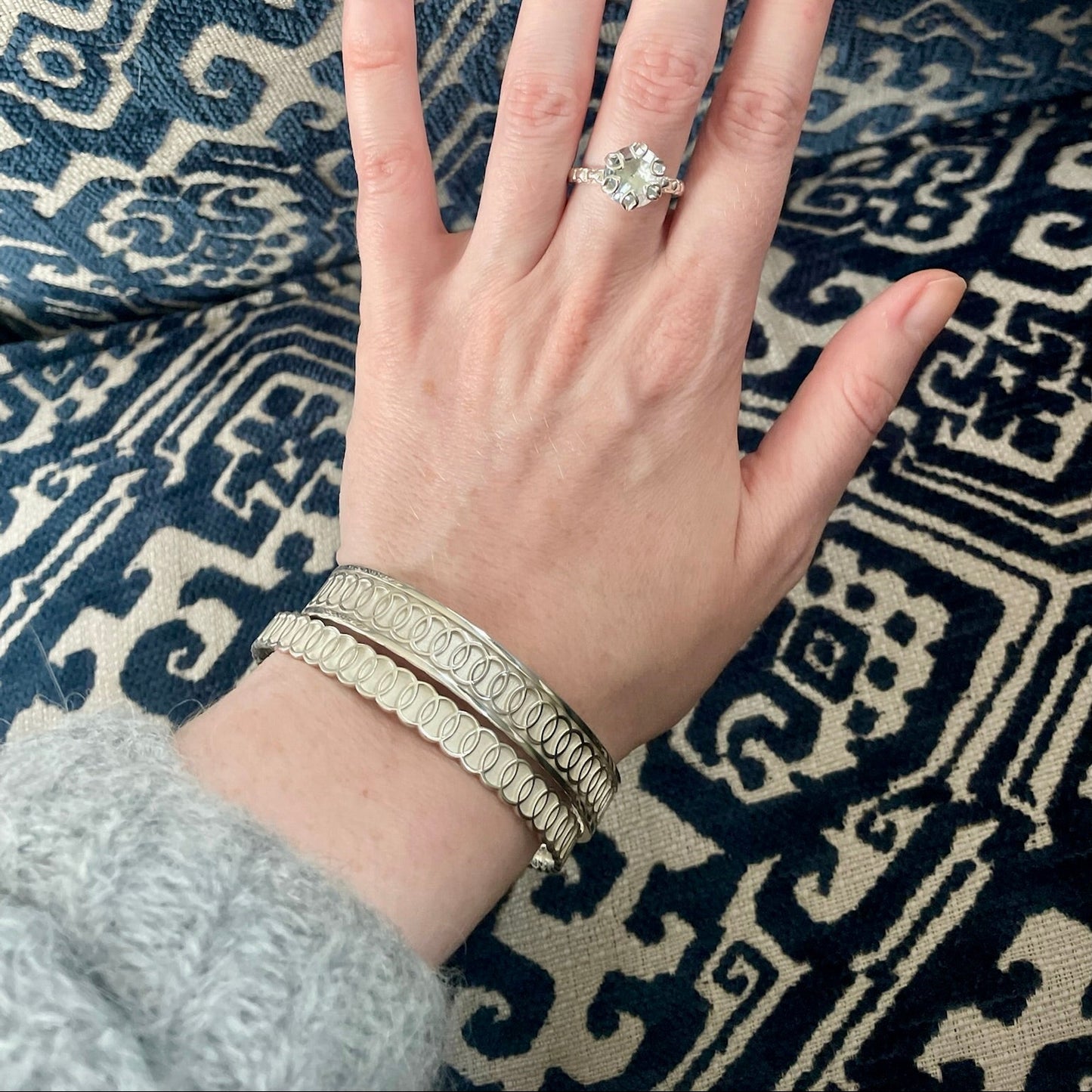 A hand wearing a silver ring with a clear round gemstone, styled with stacked silver bracelets, resting on a navy and beige patterned textile.
