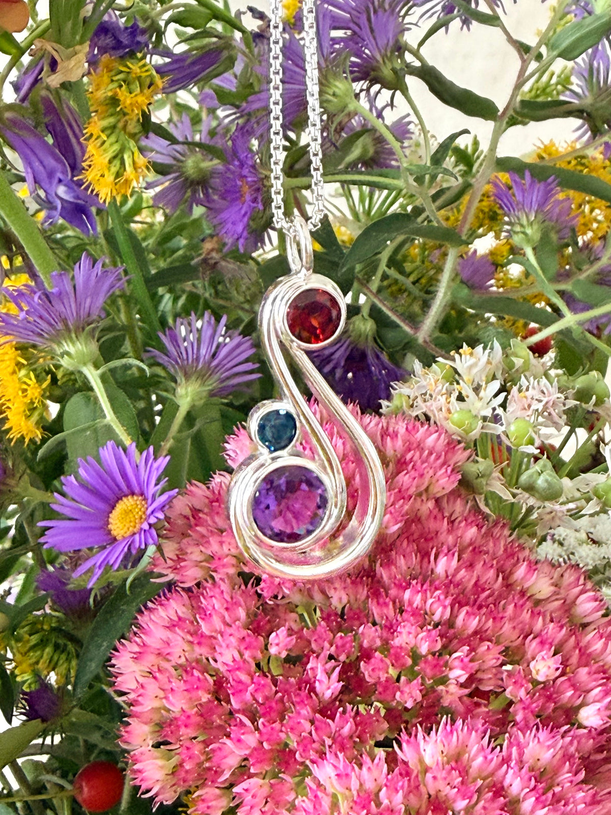 A silver pendant necklace with three gemstone accents in red, blue, and purple, displayed against a dense background of pink and purple wildflowers.