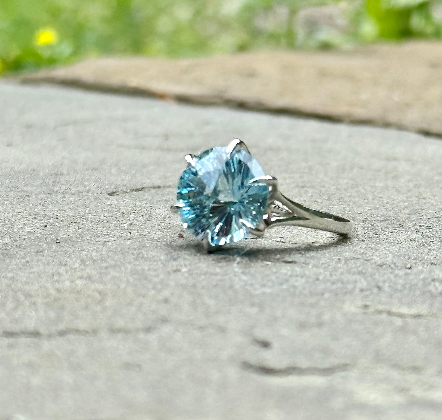 A silver ring with a large round-cut light blue gemstone, photographed outdoors.