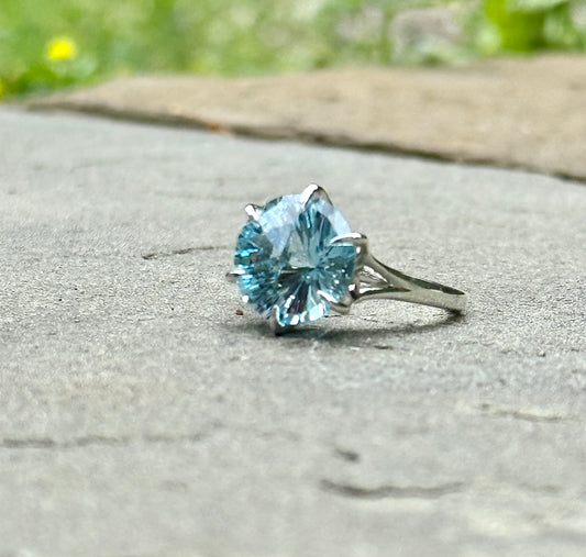 A silver ring with a large round-cut light blue gemstone, photographed outdoors.