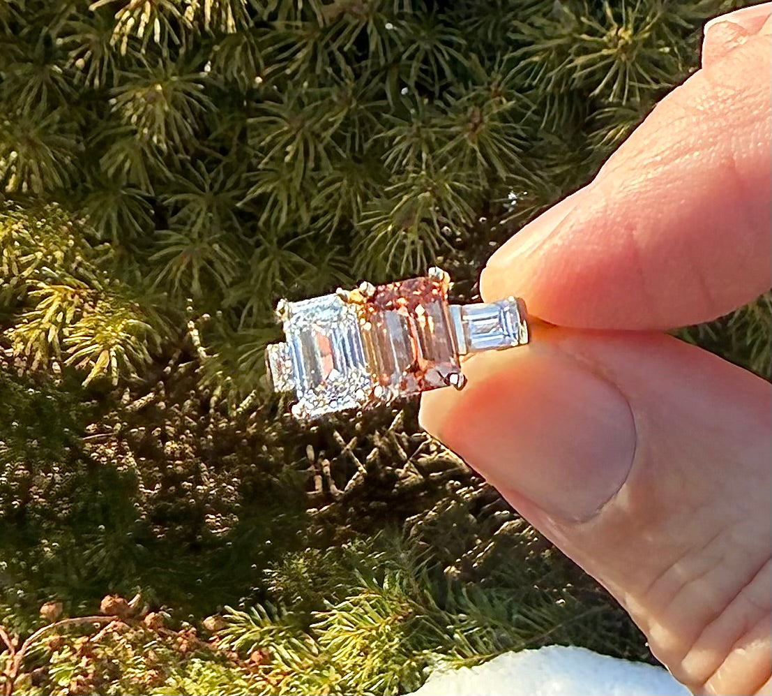 A close-up of a hand holding a rectangular emerald-cut diamond ring with baguette side stones and small round accent stones, set against a background of green pine needles.