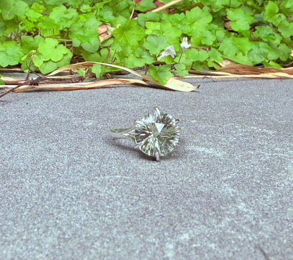 A single round brilliant-cut green amethyst ring set in silver-toned metal, resting on a gray concrete surface with green leafy plants in the background.