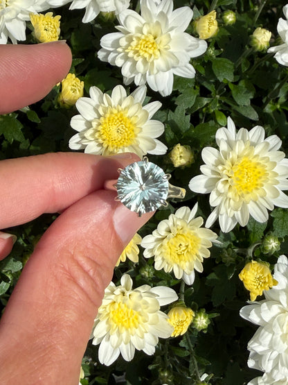 A hand holding a silver ring with a round, faceted light green gemstone, set against a background of white and yellow chrysanthemums.