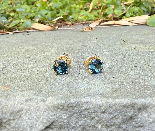 A pair of gold stud earrings with round blue-green gemstones resting on a gray stone surface outdoors with green foliage in the background.