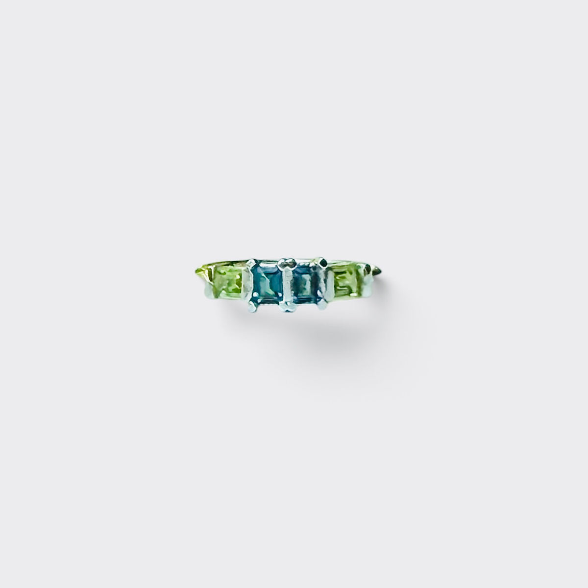 A delicate ring featuring four gemstones in green, blue, blue, and green, set in a thin silver band against a light gray background.