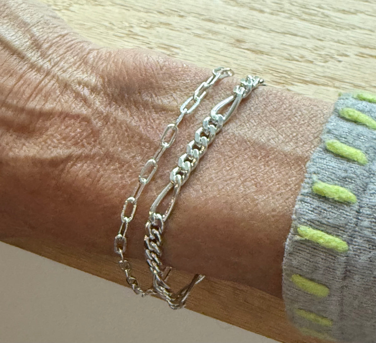 A close-up of a person's wrist wearing two silver chain bracelets, one with elongated oval links and one with curb links, paired with a gray knit sleeve featuring lime green stripes.