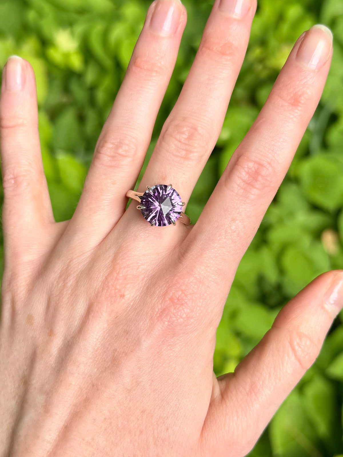 A close-up of a hand wearing a ring with a round purple faceted gemstone on a silver band, set against a blurred green background.