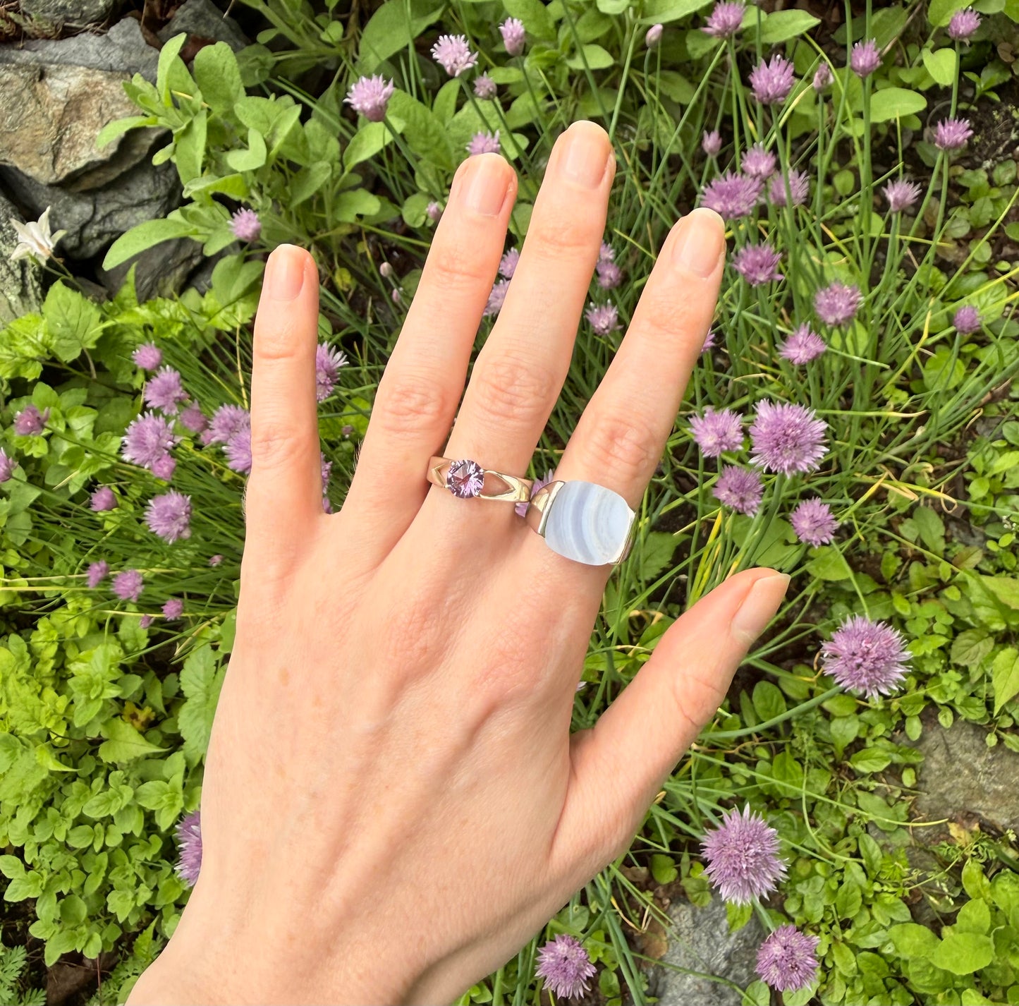 Hand wearing two rings each with a purple stone against a background of green foliage and small purple flowers
