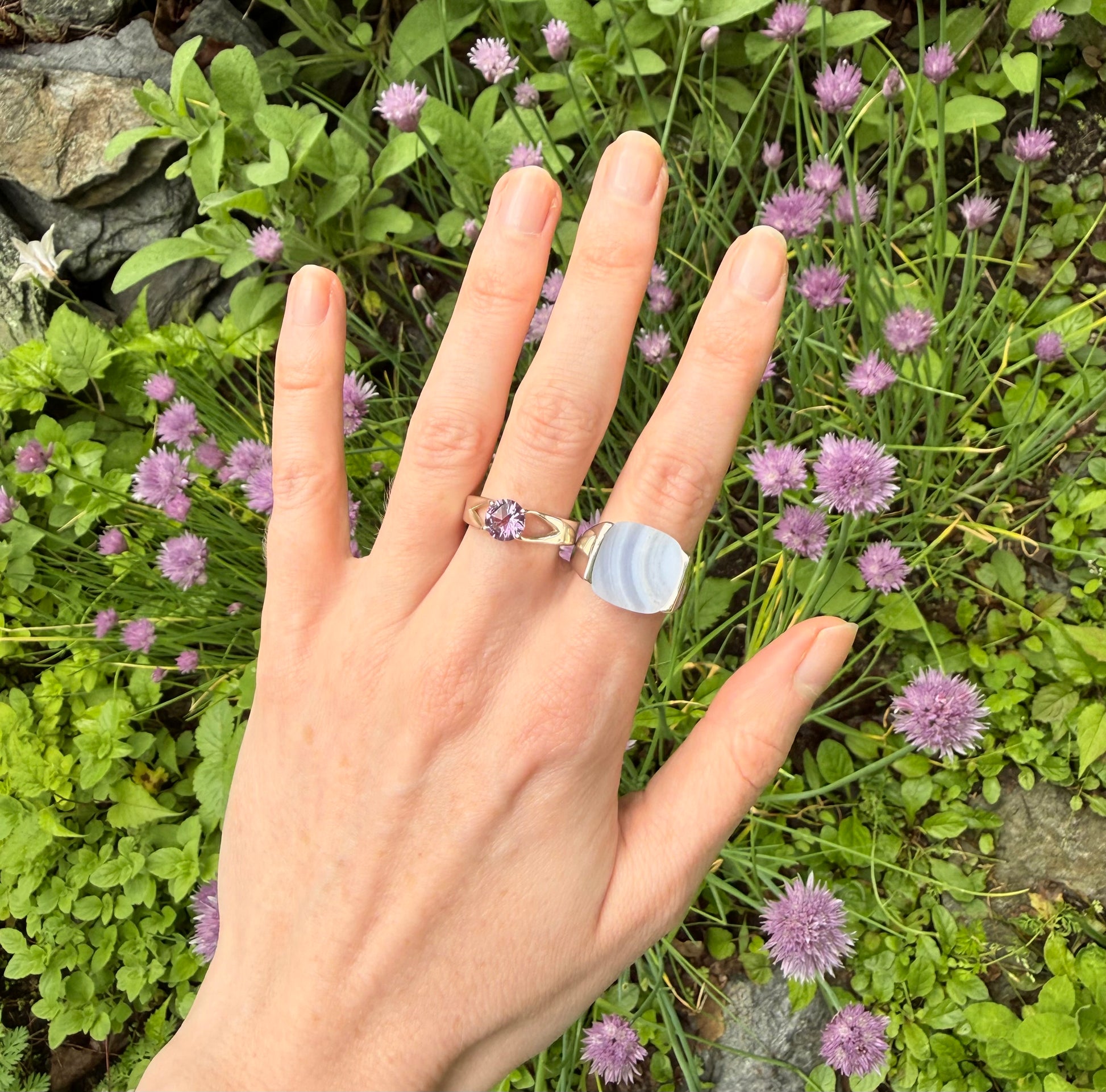 Hand wearing two rings each with a purple stone against a background of green foliage and small purple flowers