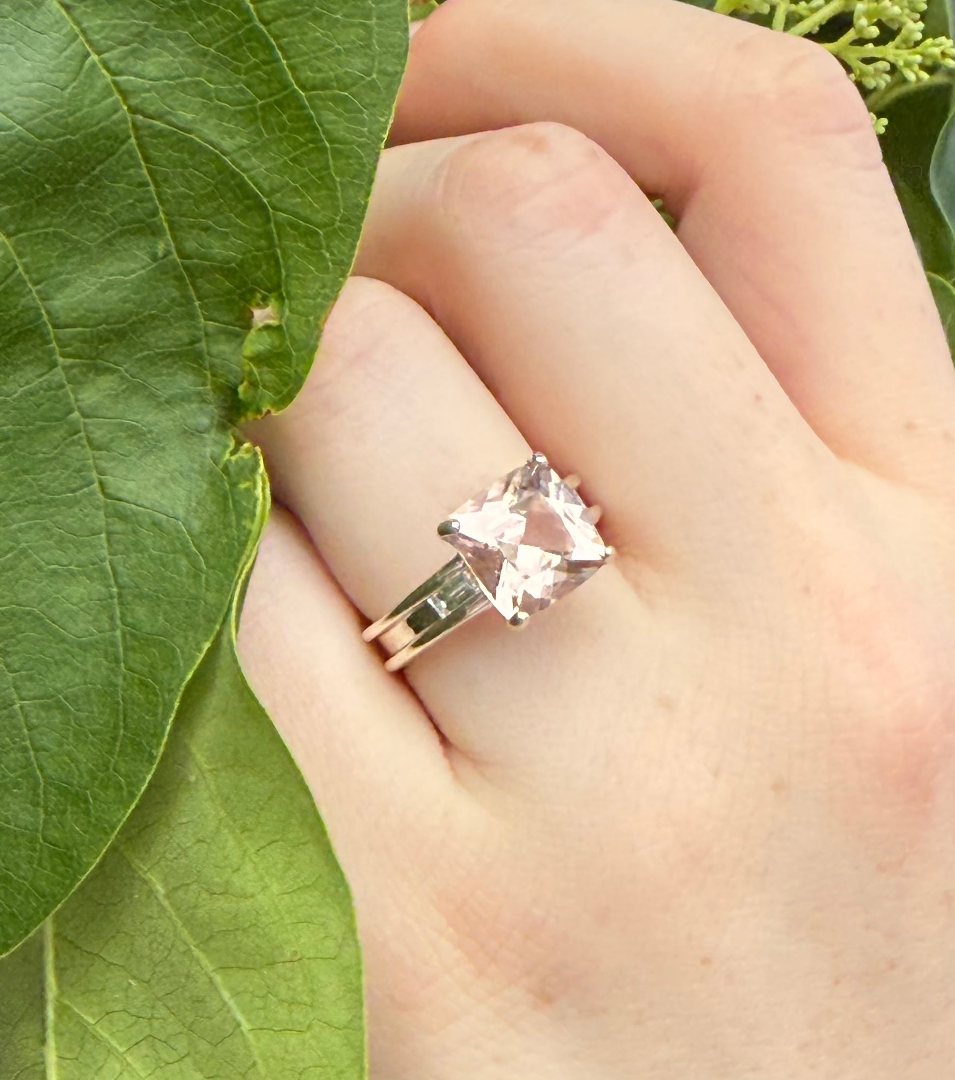 Hand wearing a ring with a pink gemstone against a green leaf background