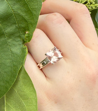 Hand wearing a ring with a pink gemstone against a green leaf background