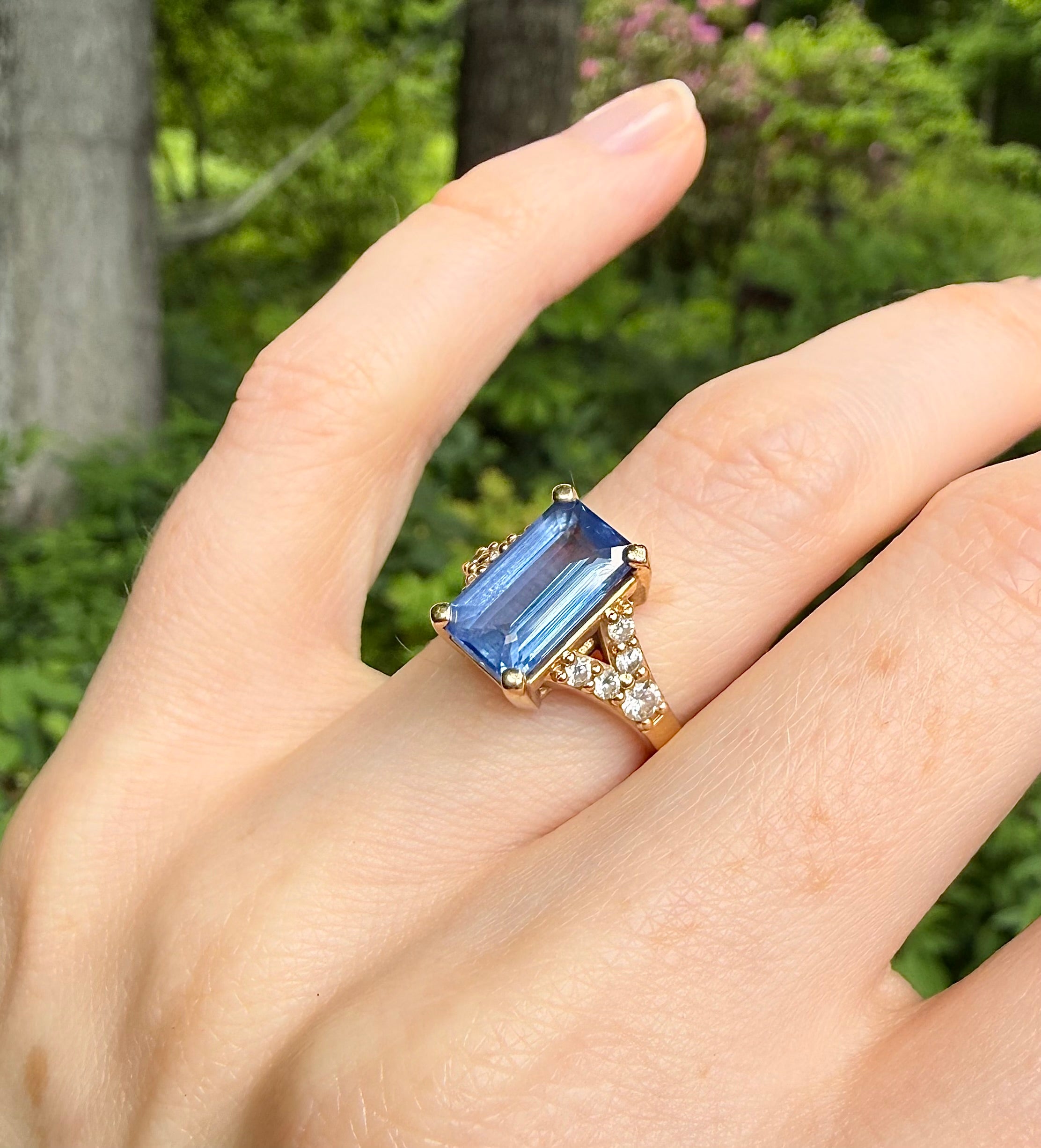 Hand wearing a ring with a blue sapphire gemstone against a green outdoor background