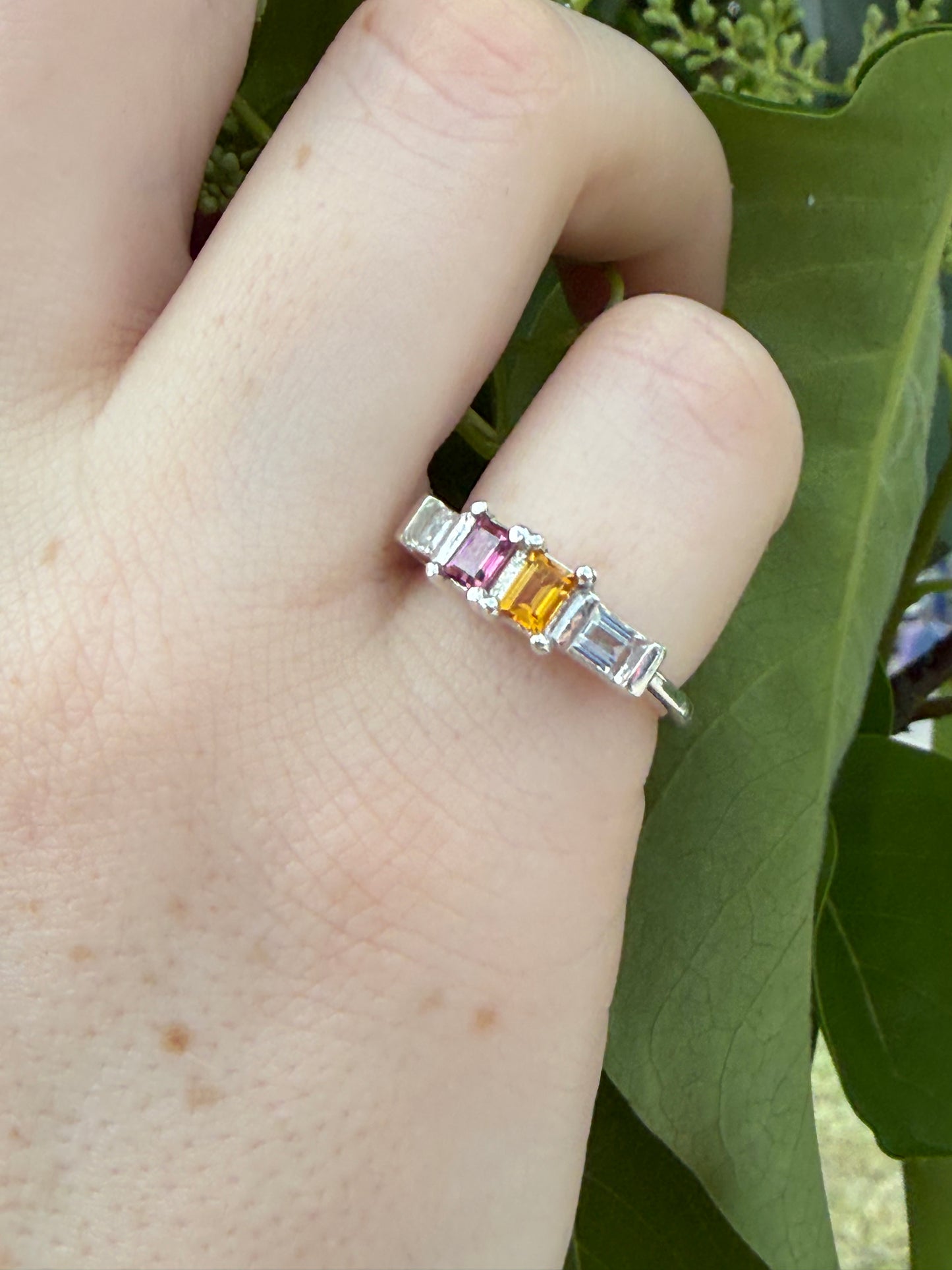 Silver ring with multicolored gemstones on a person's finger against a green leafy background