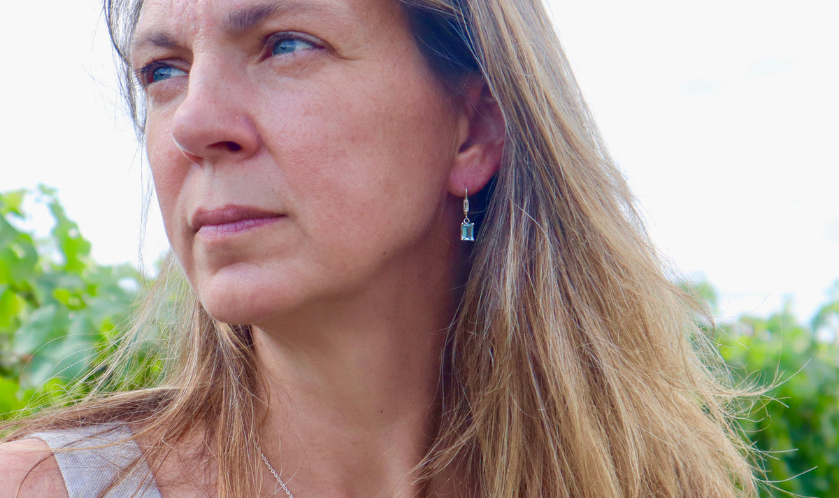 A close-up lifestyle shot of a woman with long blonde hair wearing a small, dangling blue earring with a silver-toned hook, set against a soft-focus background of green foliage and bright sky.