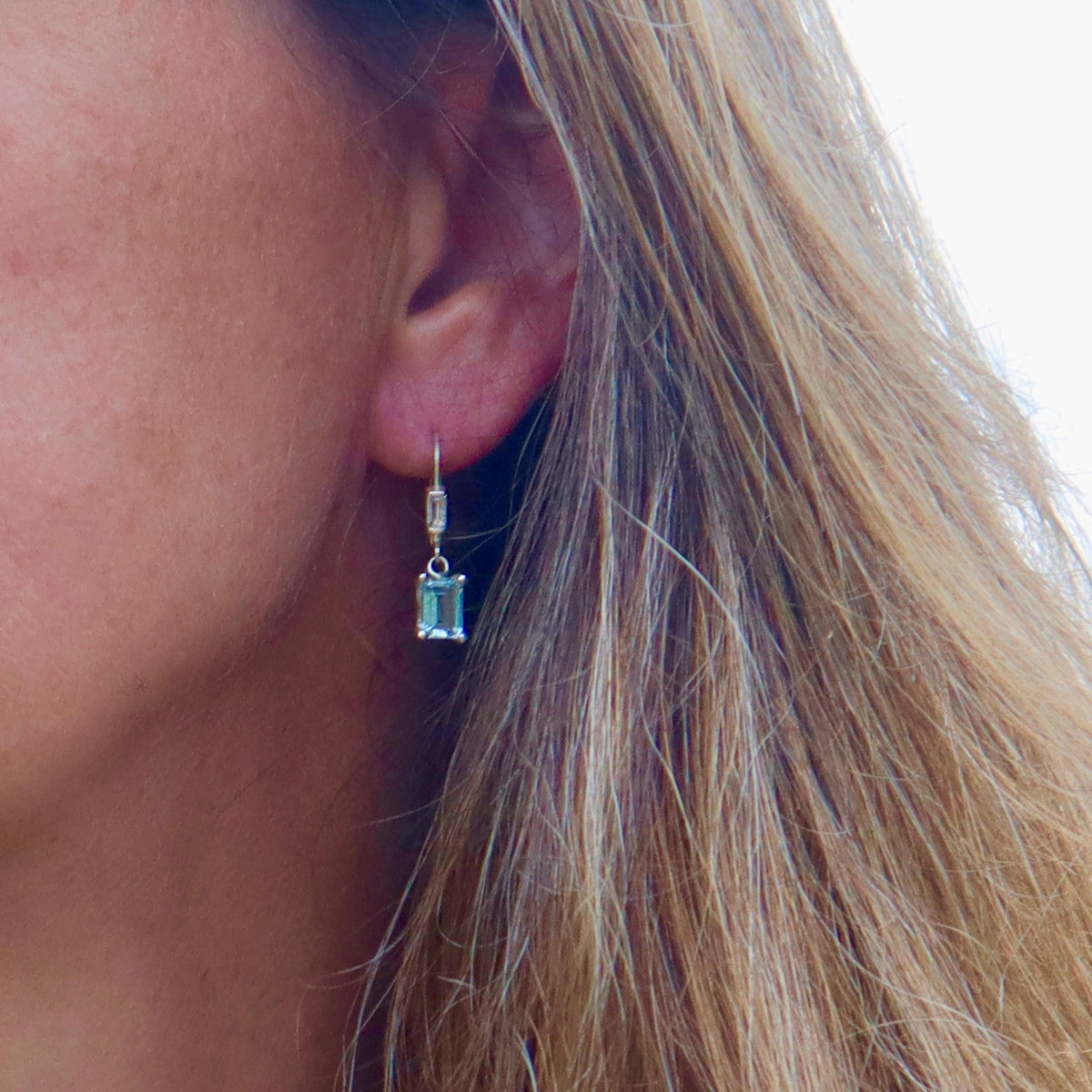 A close-up of a woman's ear wearing a silver drop earring with a rectangular light blue gemstone.