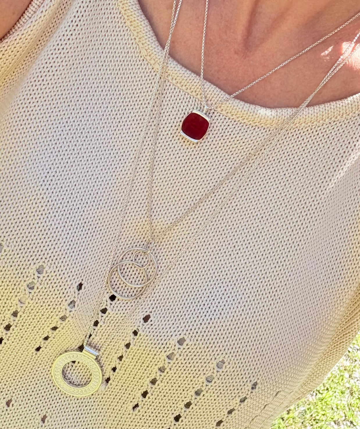 A close-up of a person wearing layered silver necklaces over a cream-colored knit top. The necklaces include a square red pendant, a circular pendant, and a double-ring pendant.