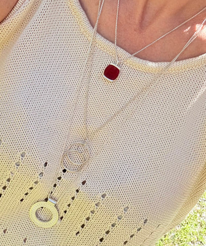 A close-up of a person wearing layered silver necklaces over a cream-colored knit top. The necklaces include a square red pendant, a circular pendant, and a double-ring pendant.