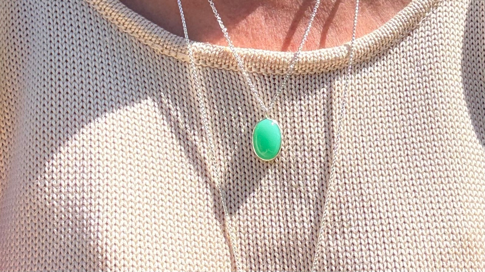 A close-up of a person wearing a delicate silver necklace with an oval light green pendant, layered over a cream-colored knit top.