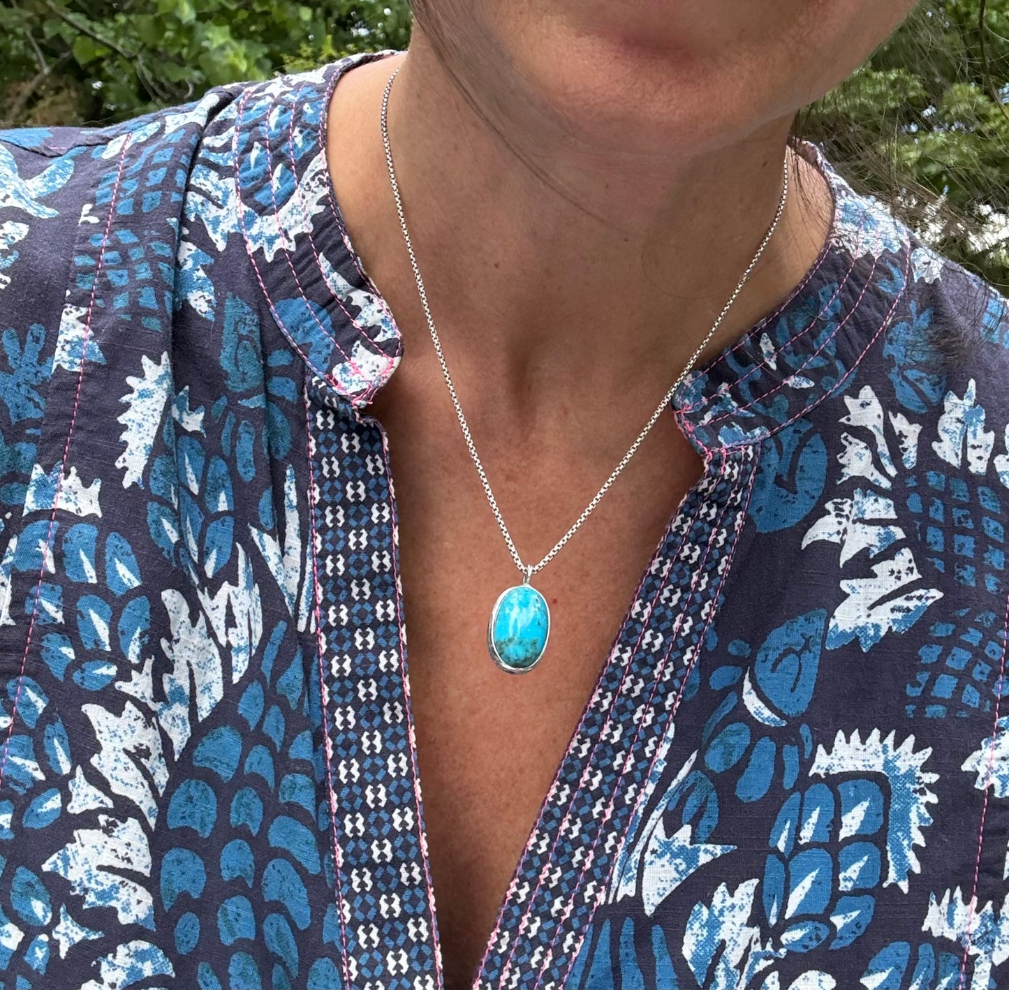 A close-up of a person wearing a silver chain necklace with an oval turquoise-blue stone pendant, paired with a blue and white patterned blouse.
