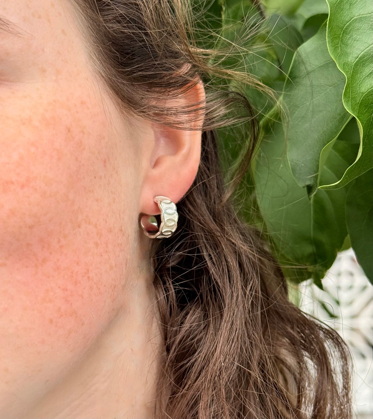 Close-up of an ear wearing a silver hoop earring with green leaves in the background
