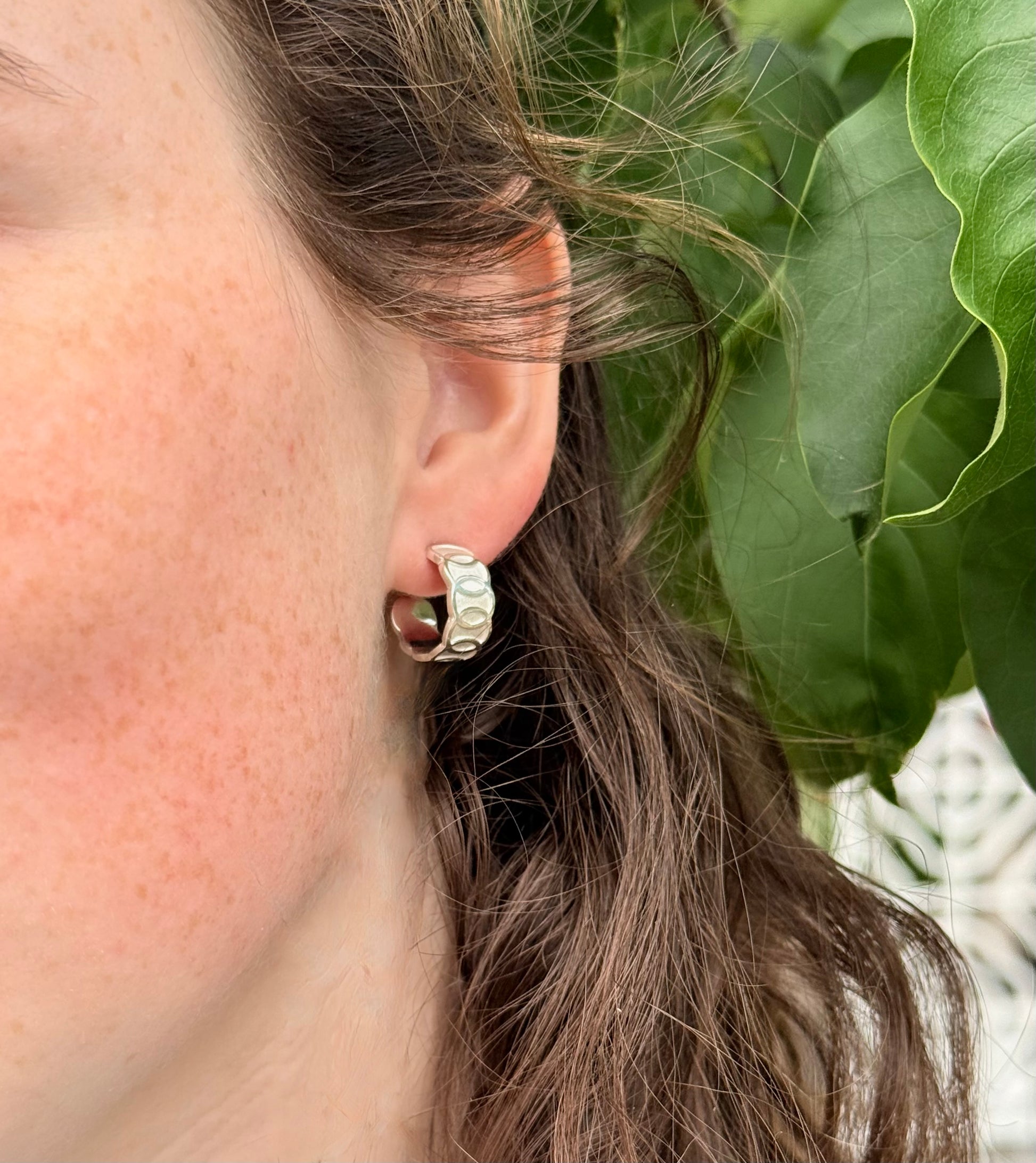 Close-up of an ear wearing a silver hoop earring with green leaves in the background