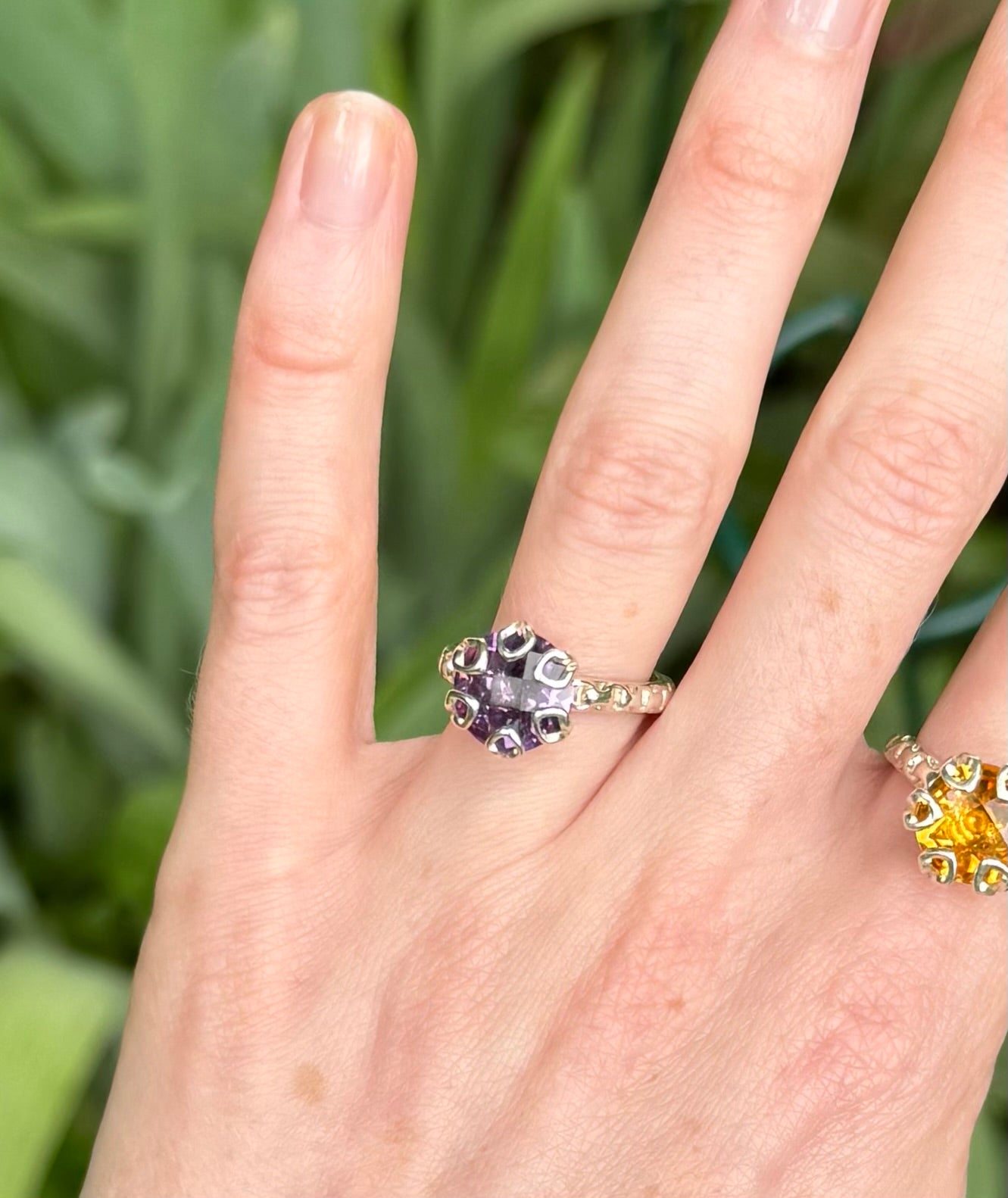 Hand wearing a decorative amethyst purple ring with green foliage in the background