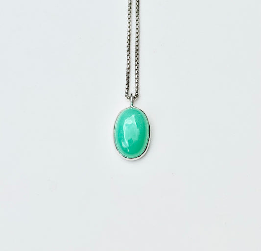 An oval-shaped light green cabochon gemstone pendant set in a silver bezel, hanging from a silver chain against a plain white background.