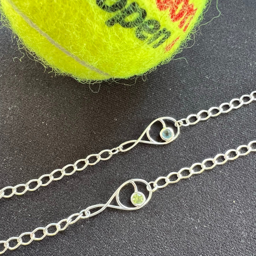 A sterling silver tennis racquet charm bracelet with a green gemstone accent, displayed next to a yellow tennis ball.
