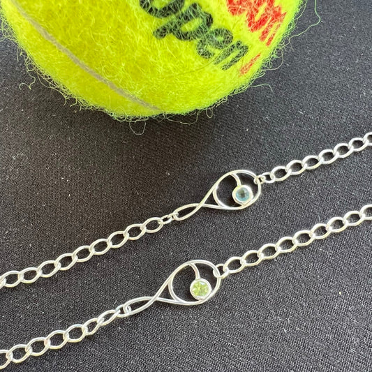 A sterling silver tennis racquet charm bracelet with a green gemstone accent, displayed next to a yellow tennis ball.